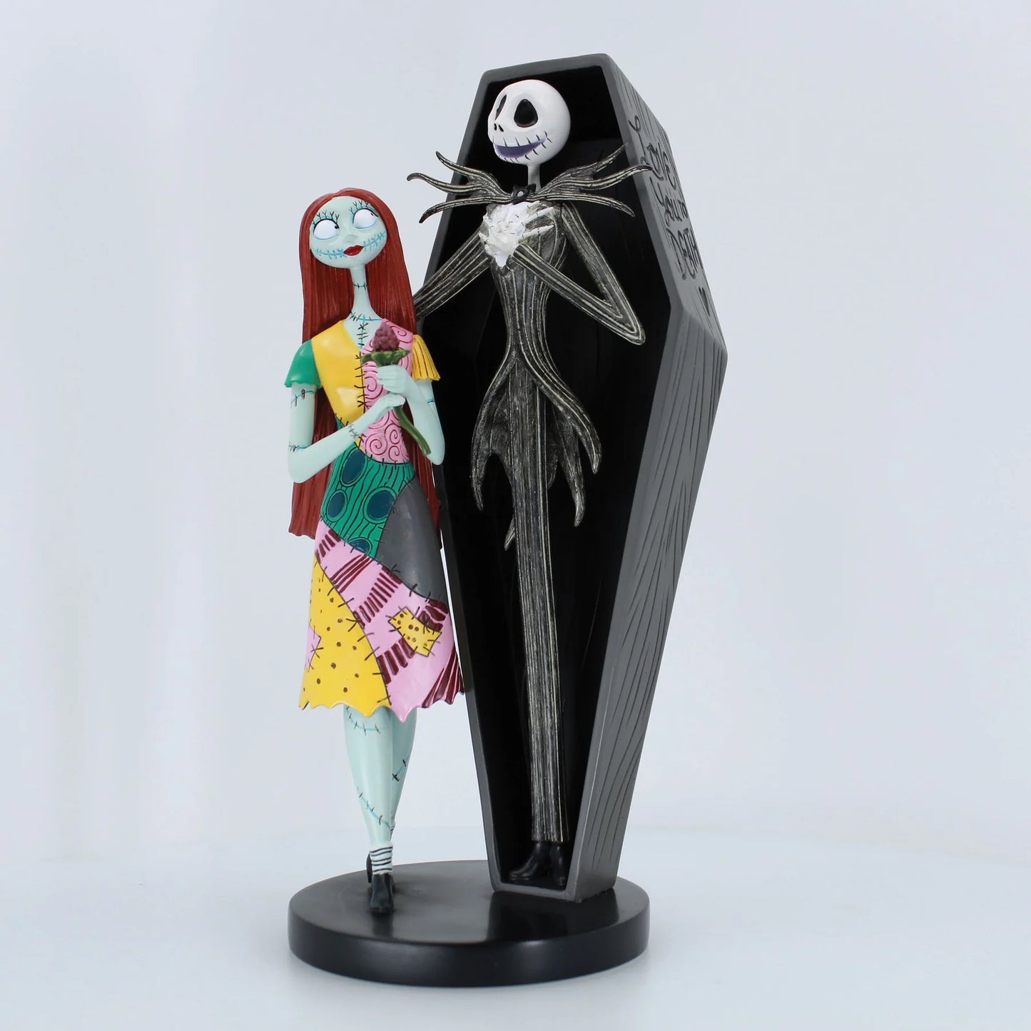 A Jack Skellington and Sally figurine set on a black base, featuring Sally in a colorful patchwork dress next to Jack inside a black coffin-shaped display stand.