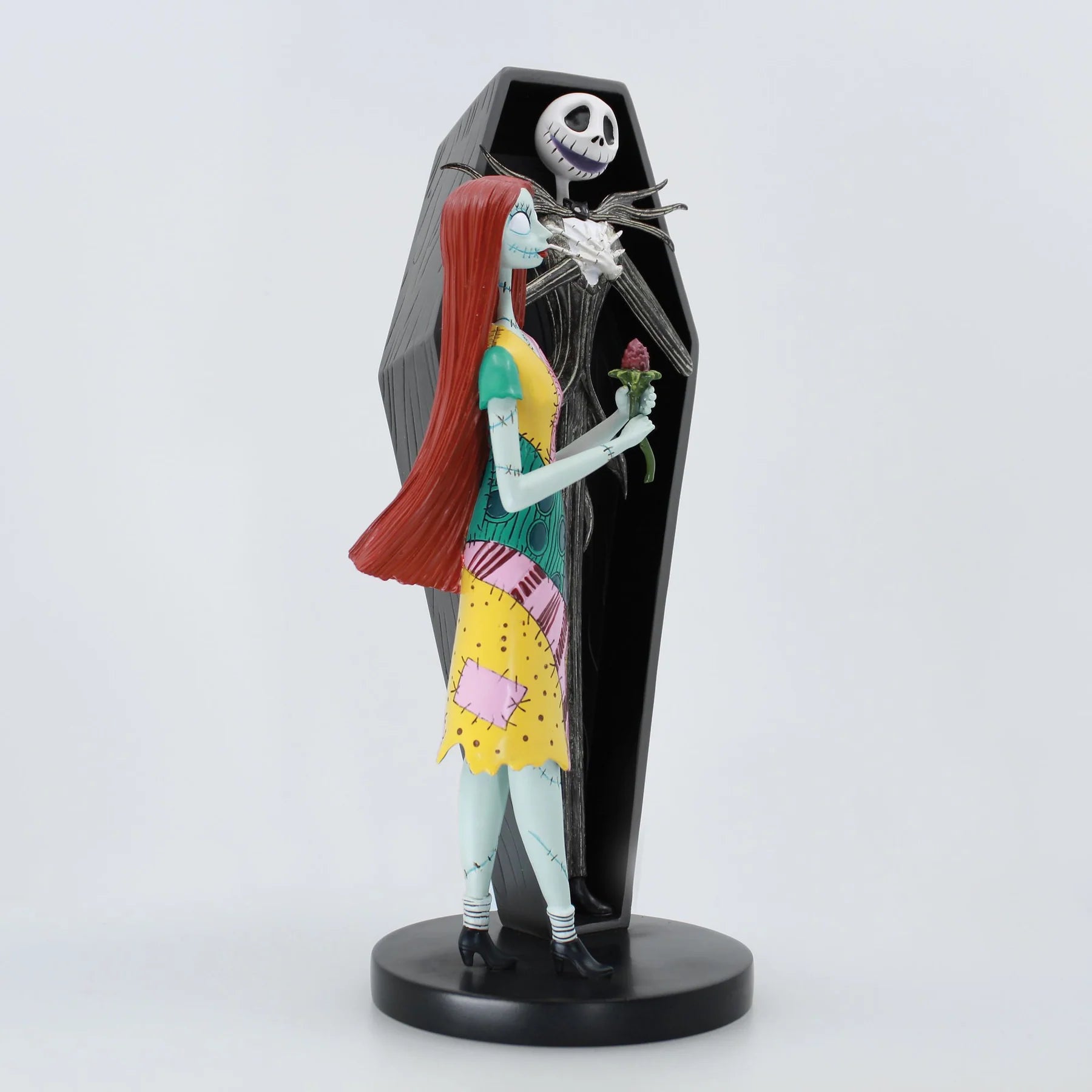 A Jack Skellington and Sally figurine from The Nightmare Before Christmas, standing in front of a black coffin-shaped backdrop on a round black base. Sally has long red hair, pale blue skin, and wears a yellow, pink, and green patchwork dress, holding a small red rose.