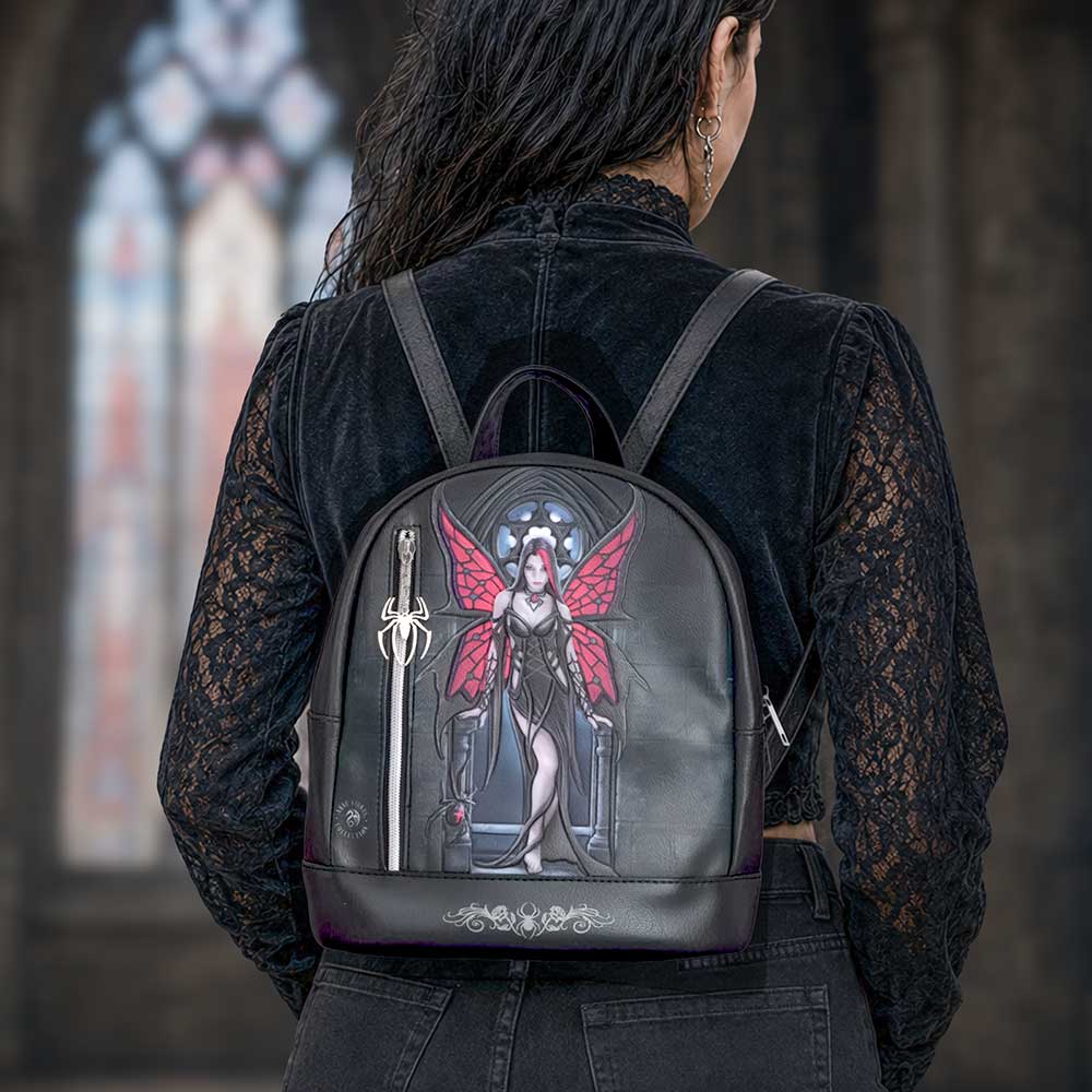 Aracnafaria Backpack by Anne Stokes displayed as a gothic fantasy accessory with a detailed fantasy illustration of a winged fairy on the front. The person is wearing a black lace long-sleeve top and dark jeans, standing in front of a blurred gothic-style stone archway with a stained glass window.