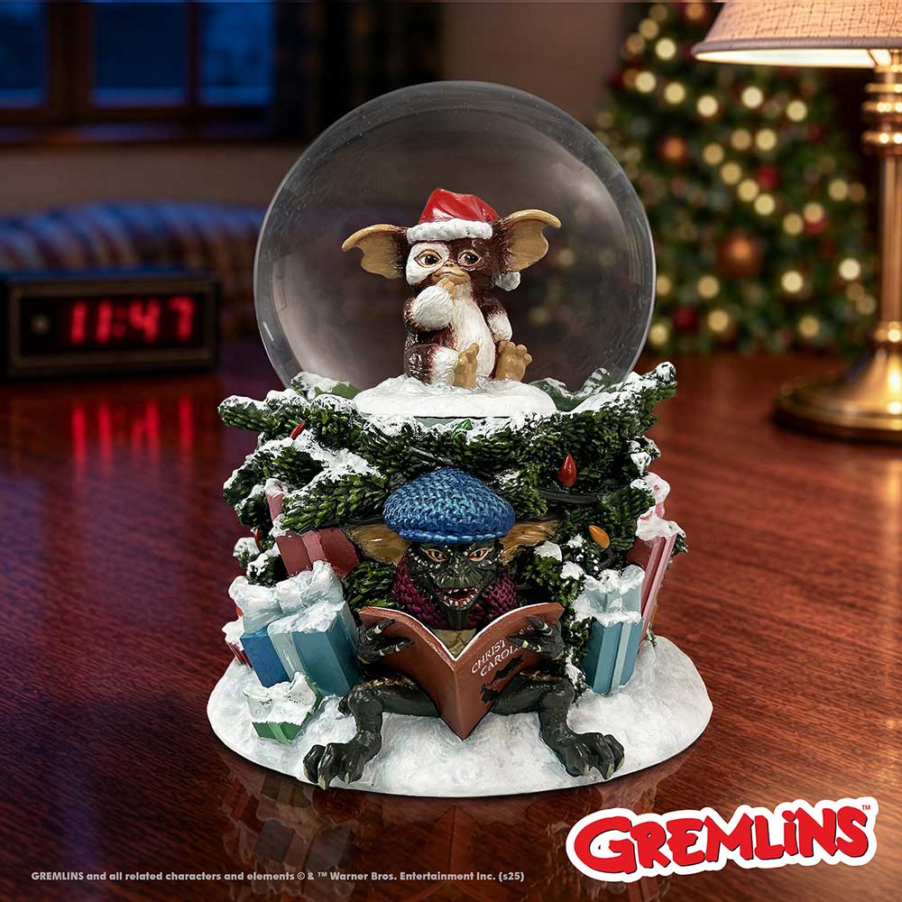 A Gremlins-themed snow globe figurine on display. The globe contains a small gremlin wearing a Santa hat, surrounded by snowy greenery, wrapped gifts, and a gremlin reading a book at the base. 